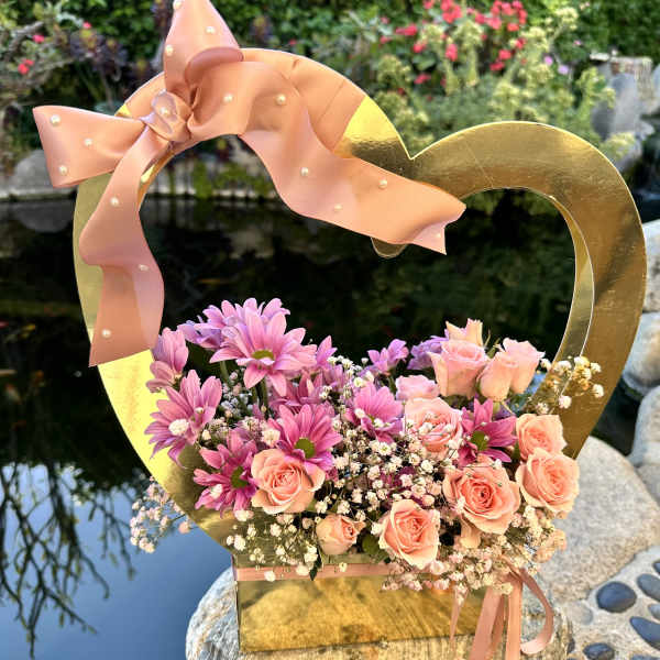 Pink roses and daisies arranged in a gold heart-shaped box with a ribbon bow.