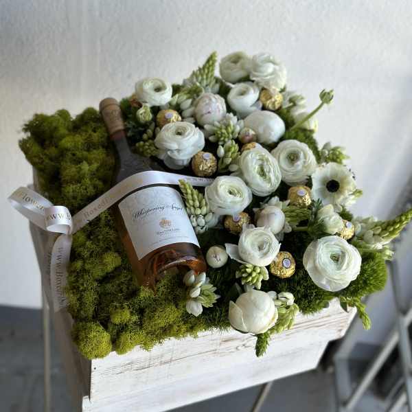 White flowers and chocolates arranged around a wine bottle in a moss-covered box