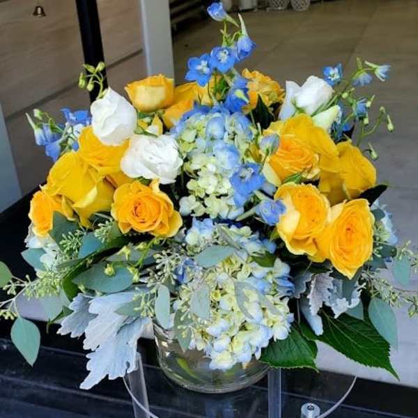 Low glass vase of yellow roses, white blooms, and blue hydrangea and delphinium