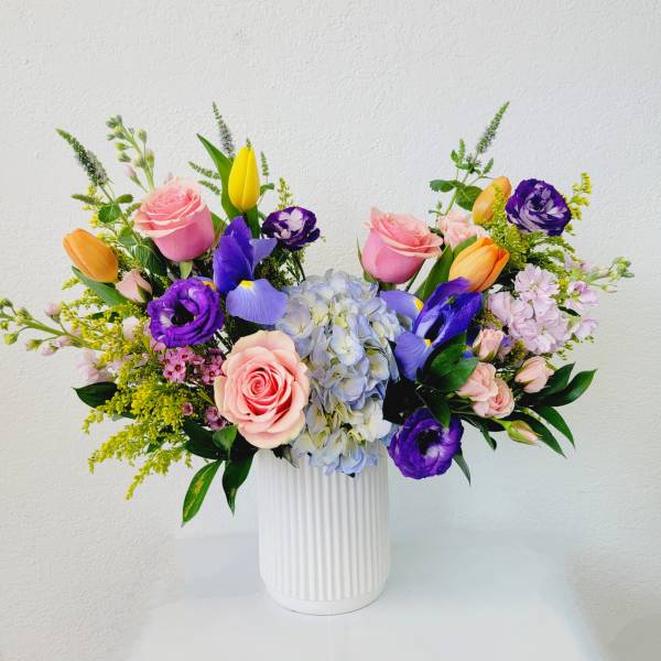 Mixed bouquet of pink roses, purple blooms, and a blue hydrangea in a white vase