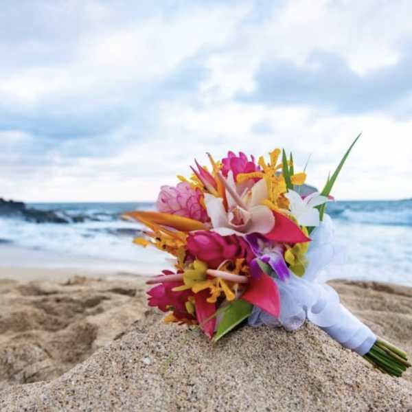 Bright tropical hand-tied bouquet with pink, orange, and yellow flowers resting on sand by the ocean