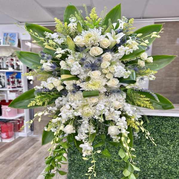Tall standing spray of white roses, lilies, orchids, and mums with large green leaves and touches of yellow
