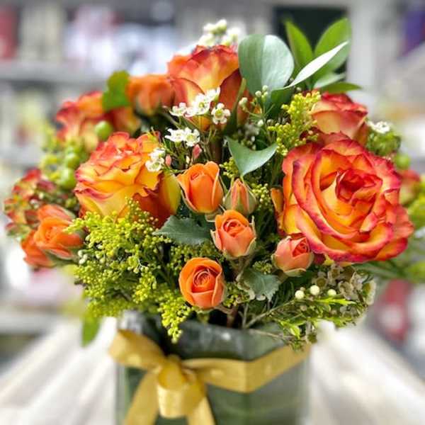Orange and yellow roses with small white flowers in a leaf-lined square glass vase with a gold ribbon bow.