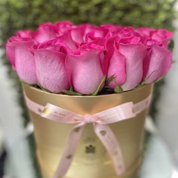 Bright pink roses arranged in a round gold hatbox with a tied pink ribbon.