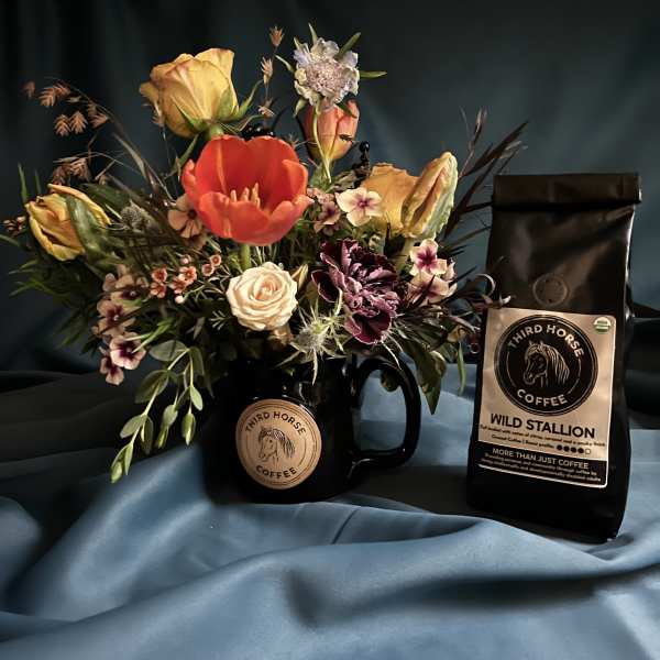 Colorful mixed flower arrangement in a black coffee mug beside a matching bag of coffee.
