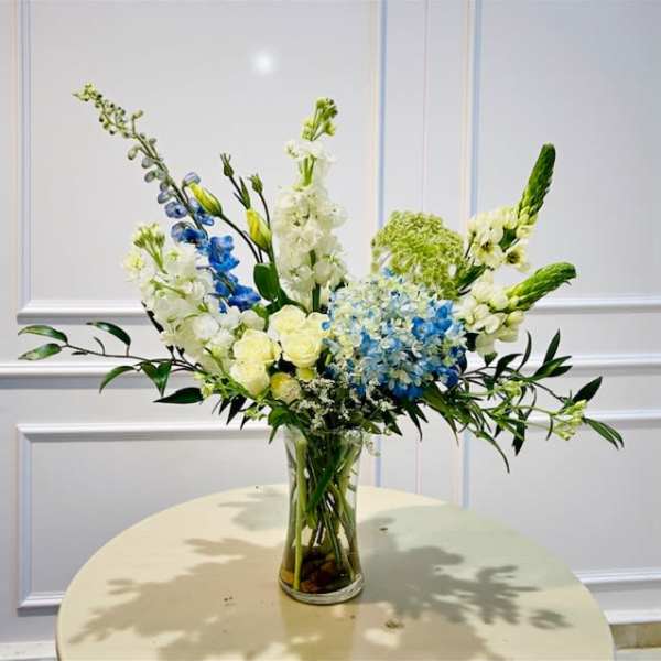 Tall floral arrangement in a clear glass vase with blue, white, and pale yellow blooms