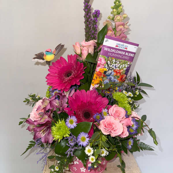 Mixed bouquet with pink gerbera daisies, roses, and purple flowers in a pink container