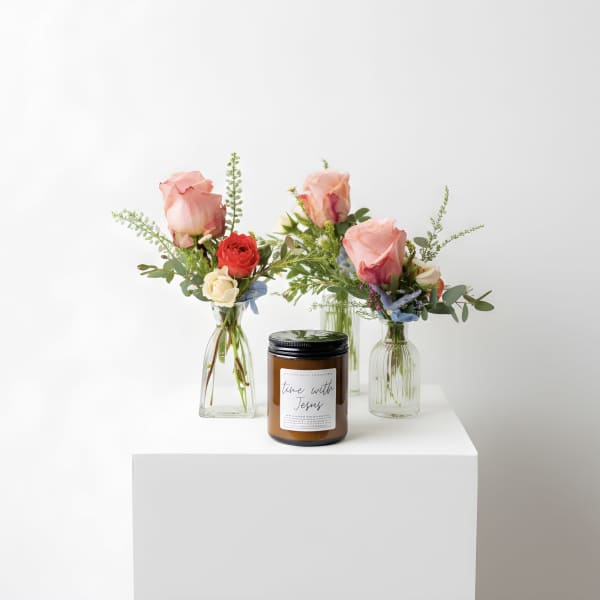 Three small bud vases with pink and red mixed flowers arranged around an amber jar candle on a white pedestal.