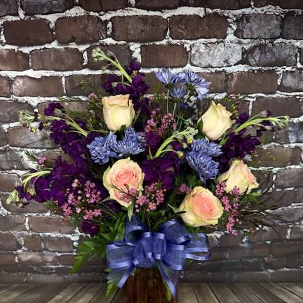 Bouquet of cream roses, purple flowers, and blue blooms in a glass vase with a ribbon