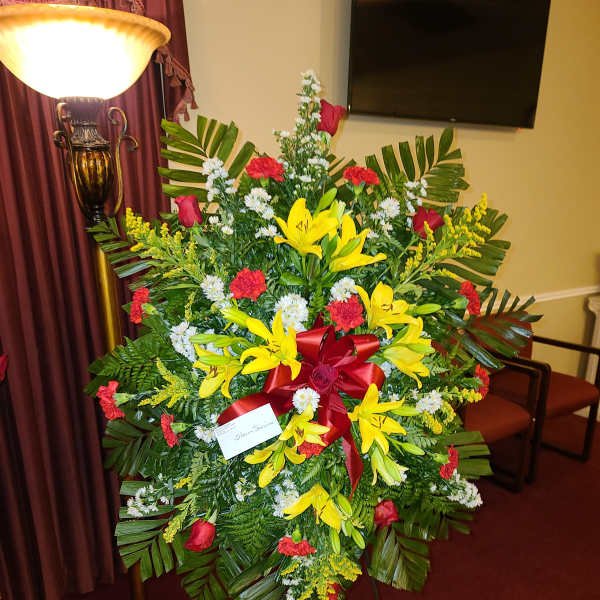 Large standing floral spray with yellow lilies, red roses, and a red ribbon