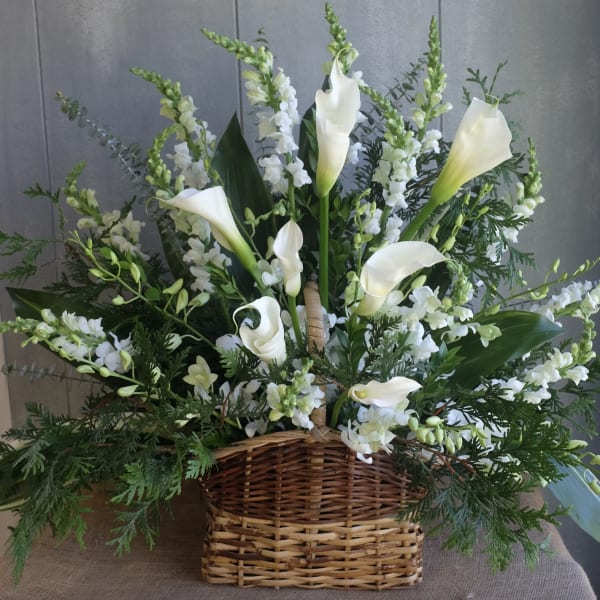 Tall arrangement of white calla lilies and other white flowers with greenery in a woven basket
