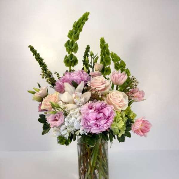 Tall pastel arrangement of pink peonies and roses with green Bells of Ireland in a clear glass vase