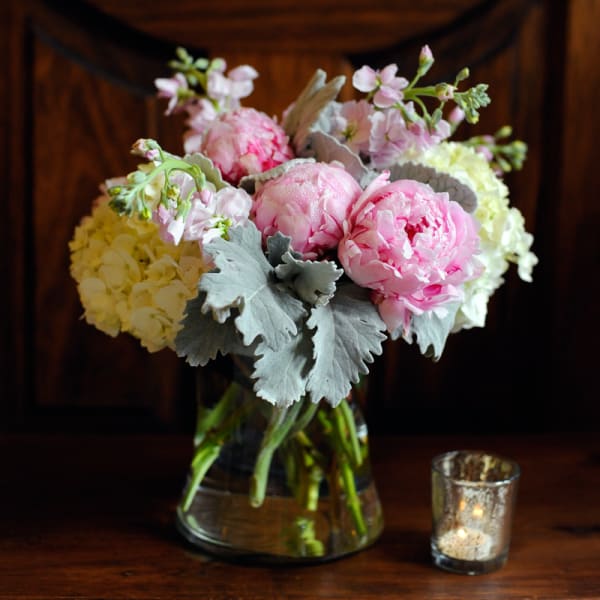 Pink peonies and pale hydrangeas in a glass vase with a small candle beside it