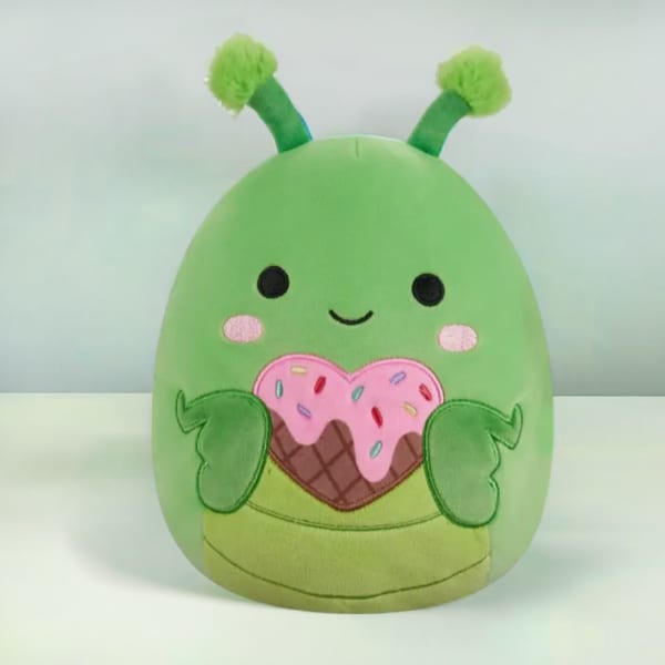 Green plush character with antennae holding a pink sprinkled heart-shaped treat.