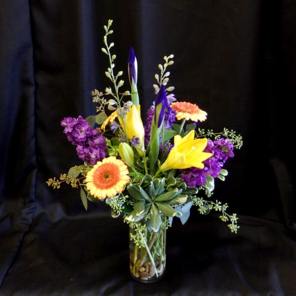 Mixed bouquet with yellow lilies, purple flowers, and orange gerbera daisies in a glass vase
