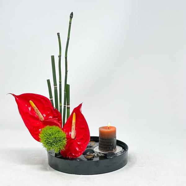 Red anthuriums with bamboo stalks and a lit candle in a black tray