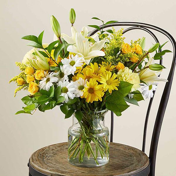 Yellow and white lilies, roses, and daisies arranged in a clear glass vase on a wooden chair