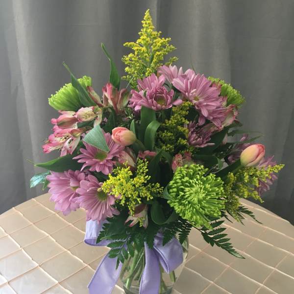 Mixed bouquet of lavender daisies, green mums, and pink blooms in a clear vase with a lavender ribbon