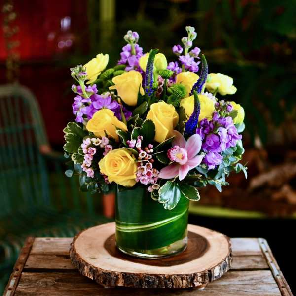 Yellow roses and purple flowers arranged in a green glass vase