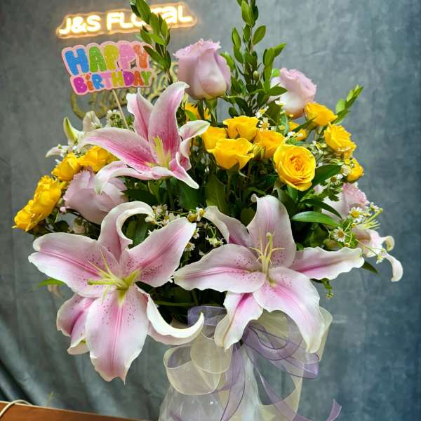 Pink lilies and yellow roses in a white vase with a birthday sign