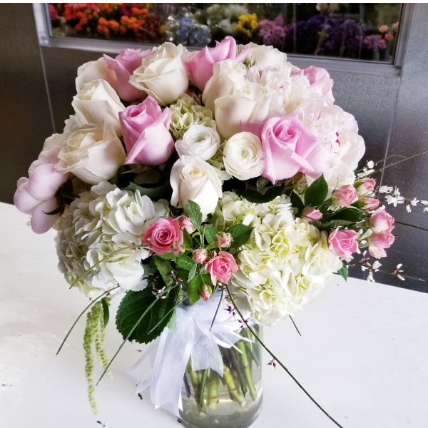 Round arrangement of white and blush pink roses with white hydrangeas in a clear glass vase with a white bow.