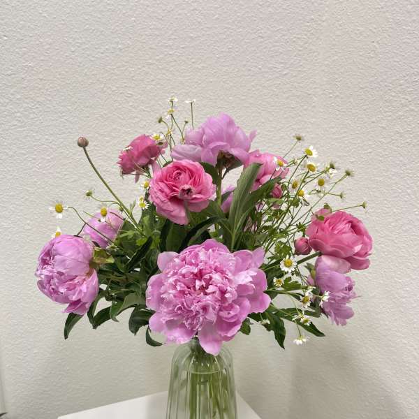 Pink peonies and roses with small white daisies arranged in a ribbed glass vase on a white pedestal