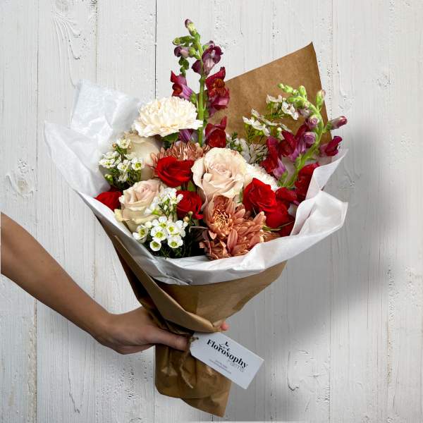 Handheld bouquet of red, blush, and white flowers wrapped in brown paper