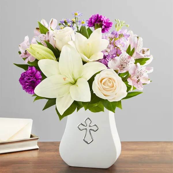 White lilies, roses, and purple flowers in a white vase with a cross emblem