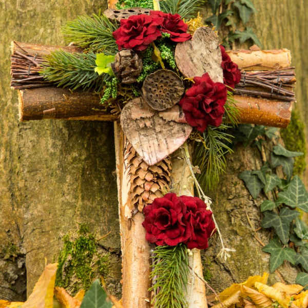 Rustic wooden cross decorated with red flowers, evergreens, bark hearts, and pinecones.
