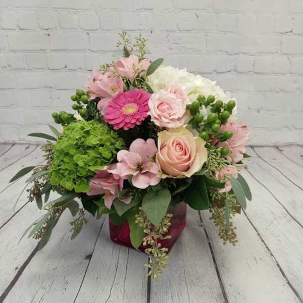 Low arrangement of pink roses and gerbera daisy with green hydrangeas in a pink glass cube vase