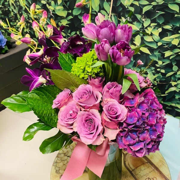 Pink roses and purple flowers arranged in a glass vase with a pink ribbon.