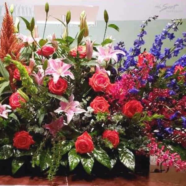 Large elongated arrangement of red roses, pink lilies, blue delphinium, and orange plumes in a low container