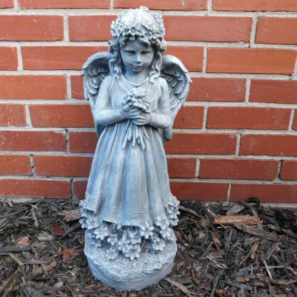 Blue-gray angel statue holding flowers in front of a brick wall