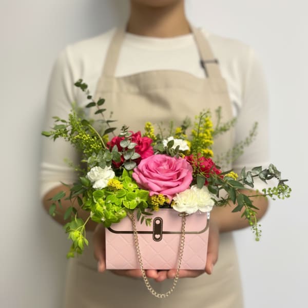 Person in an apron holding a pink purse-shaped box filled with pink, white, and green flowers.
