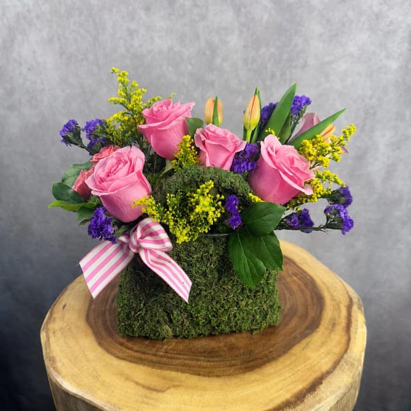 Pink roses and purple filler flowers arranged in a moss-covered box with a striped ribbon.