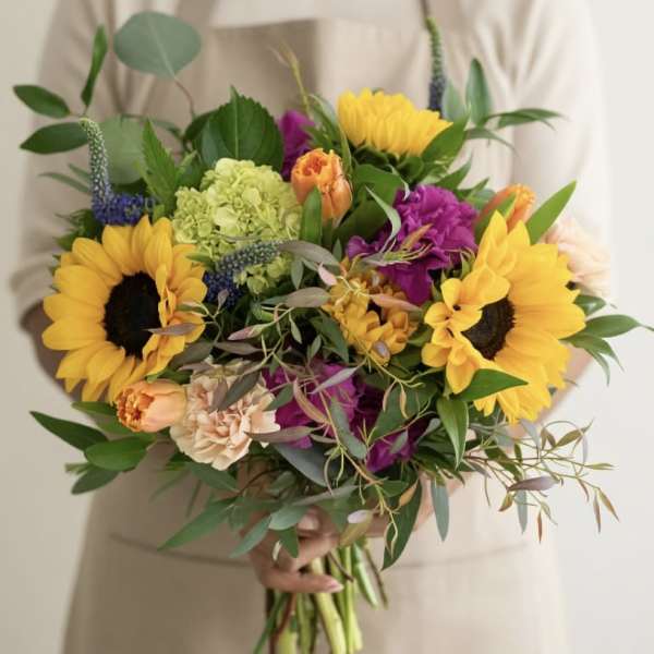 Hand-tied bouquet with bright yellow sunflowers, purple blooms, and mixed flowers held by a person in beige.
