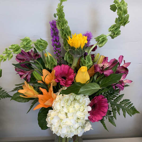 Tall mixed bouquet with white hydrangea, orange lilies, pink gerbera, yellow blooms and green Bells of Ireland in a clear vase