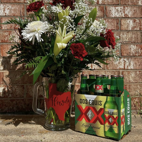 Mixed flower arrangement in a glass mug beside a six-pack of Dos Equis beer