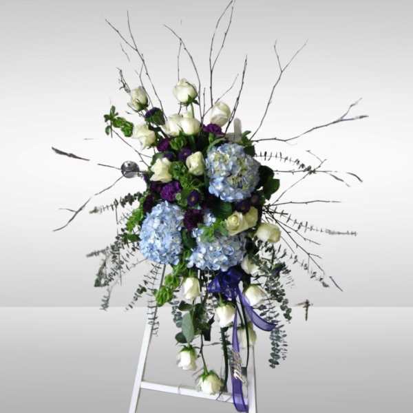 Tall floral spray with white roses, blue hydrangeas, and purple accents on an easel
