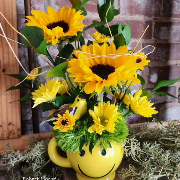 Yellow sunflower arrangement in a smiley-face mug with a small bird figurine