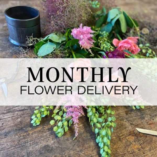 Loose flowers and greenery on a table with a black cup and monthly flower delivery text
