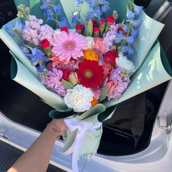 Colorful mixed bouquet with gerbera daisies, tulips, and blue flowers wrapped in mint paper