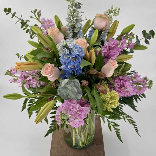 Tall pastel arrangement of pink roses, lilies, blue delphinium and stock in a clear glass vase