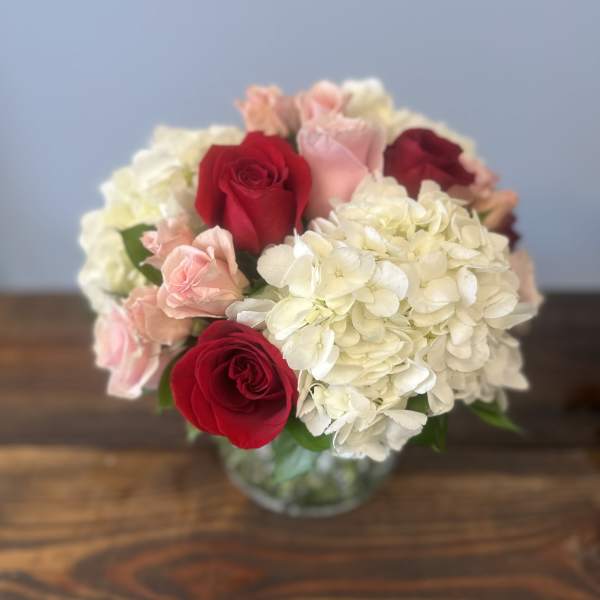 Round bouquet of red and pink roses with white hydrangeas in a clear glass vase on a wooden table
