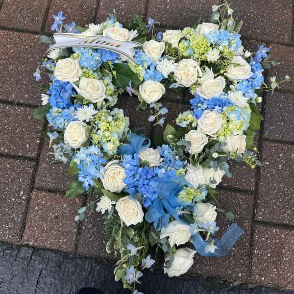 Heart-shaped wreath of white roses and blue hydrangeas with a Mom ribbon on brick pavement