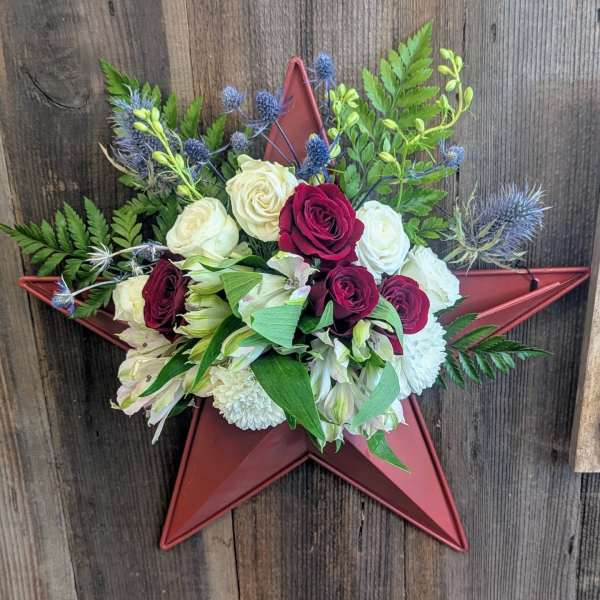 Wall arrangement of red and white roses with white mums and blue thistle on a red star.