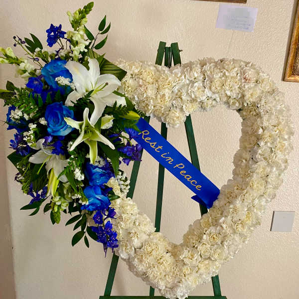 Heart-shaped white floral wreath with blue and white bouquet and a "Rest in Peace" ribbon