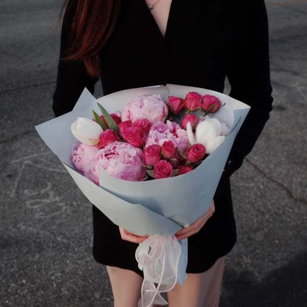 Handheld bouquet of pink peonies, roses, and white tulips wrapped in pale paper