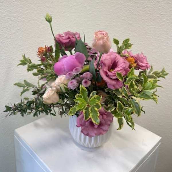 Low arrangement of pink lisianthus, roses, and mums with variegated foliage in a white ceramic vase.