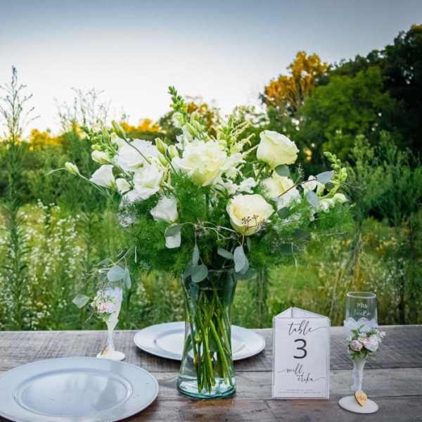White floral centerpiece in a glass vase on an outdoor table with place cards.